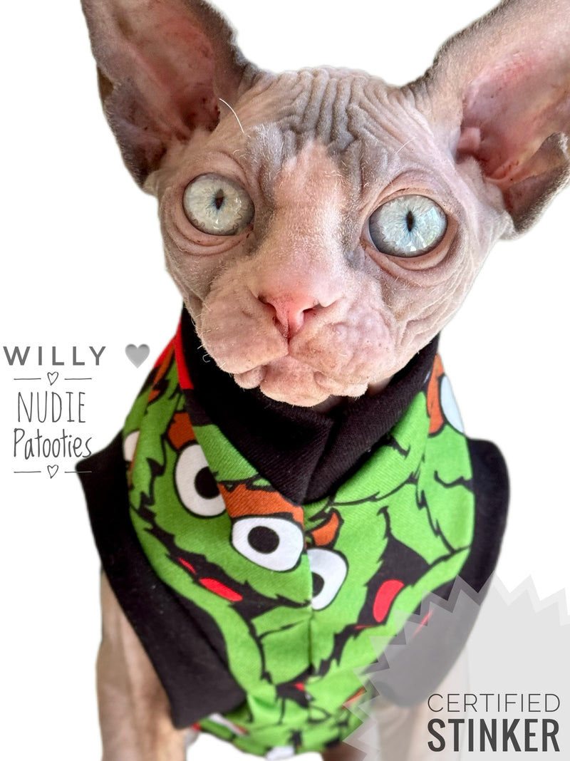 A close-up of a hairless cat wearing a green and black  sphynx cat shirt with cartoon graphics. The cat has large pink ears and blue-gray eyes, set against a plain white background with text overlays reading 'WILLY & NUDIE Patooties' and 'CERTIFIED STINKER'.